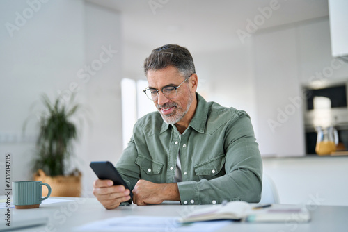 Happy mature senior man using mobile phone sitting at kitchen table. Smiling old middle aged customer holding smartphone scrolling buying online doing shopping,reading news in smartphone at home.