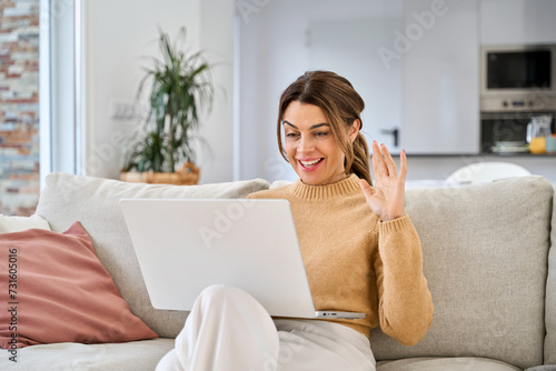 Happy mature older woman using laptop having video call sitting on couch at home. Smiling middle aged woman waving hand looking at computer chatting online relaxing on sofa in living room.