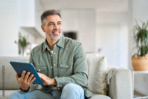 Happy middle aged man using digital tablet relaxing on couch at home. Mature male user holding tab computer holding pad technology device sitting on sofa in living room looking away. Copy space. photo