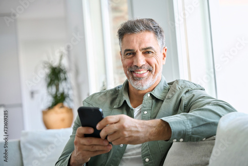 Smiling happy mature middle aged man holding cell mobile phone using smartphone sitting at home on couch, scrolling social media, checking financial apps, buying online, looking at camera. Portrait photo