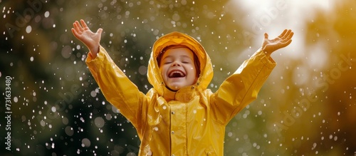 A happy little boy in a yellow raincoat smiling and gesturing with outstretched arms, enjoying the fun event of standing in the rain, surrounded by nature, art, water, and grassy surroundings.