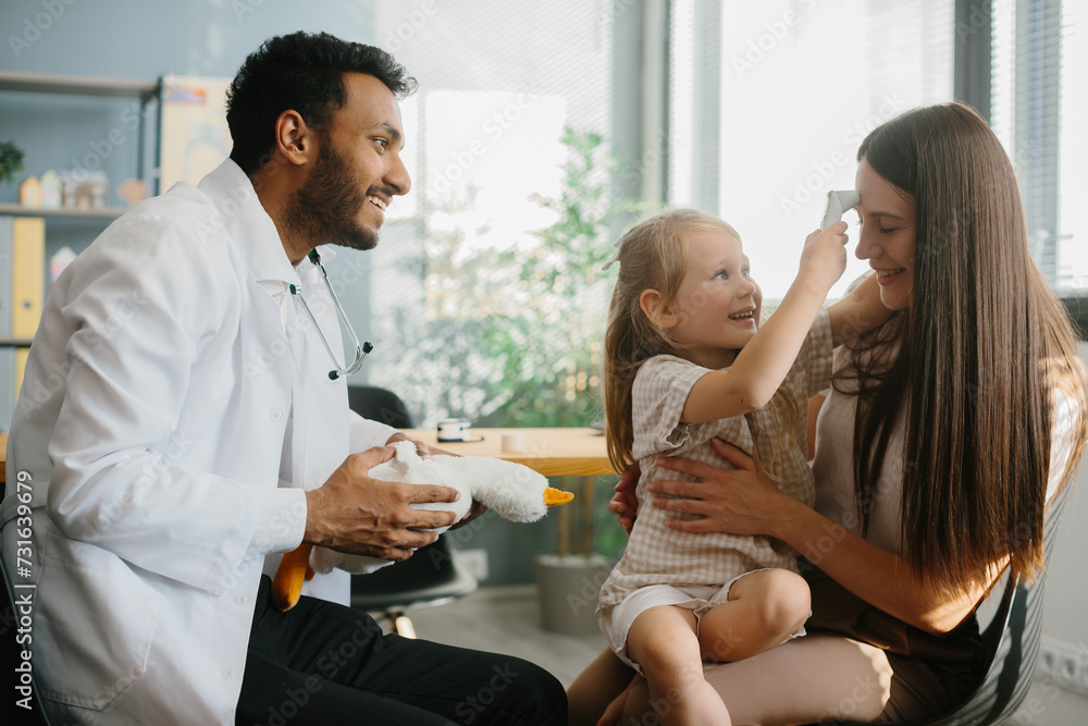 Obraz premium A young male pediatrician plays with a little girl patient in order to gain trust and conduct an examination. A young mother with a preschool-aged daughter in the doctor's office.