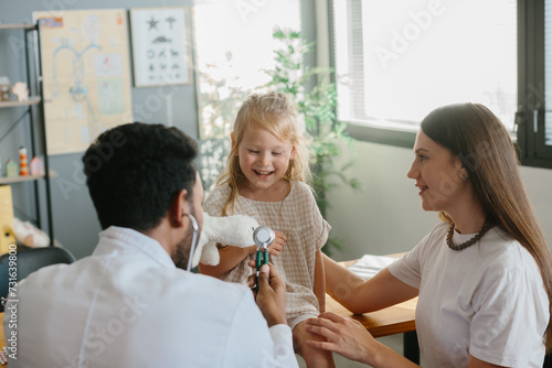 A preschool-aged girl with her mother is being examined by a pediatrician. A young man, a pulmonologist, conducts an examination using a phalendoscope. photo