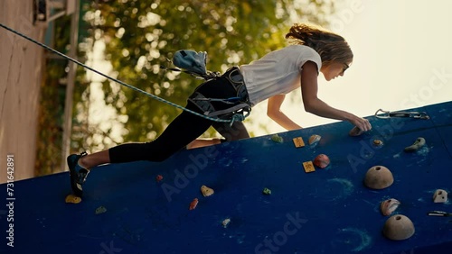 Vertical video: A blonde girl in a white T-shirt in special equipment and folding insurance climbs up a blue climbing wall in the summer. Blonde girl climber trains ska climbing and climbs up a blue photo