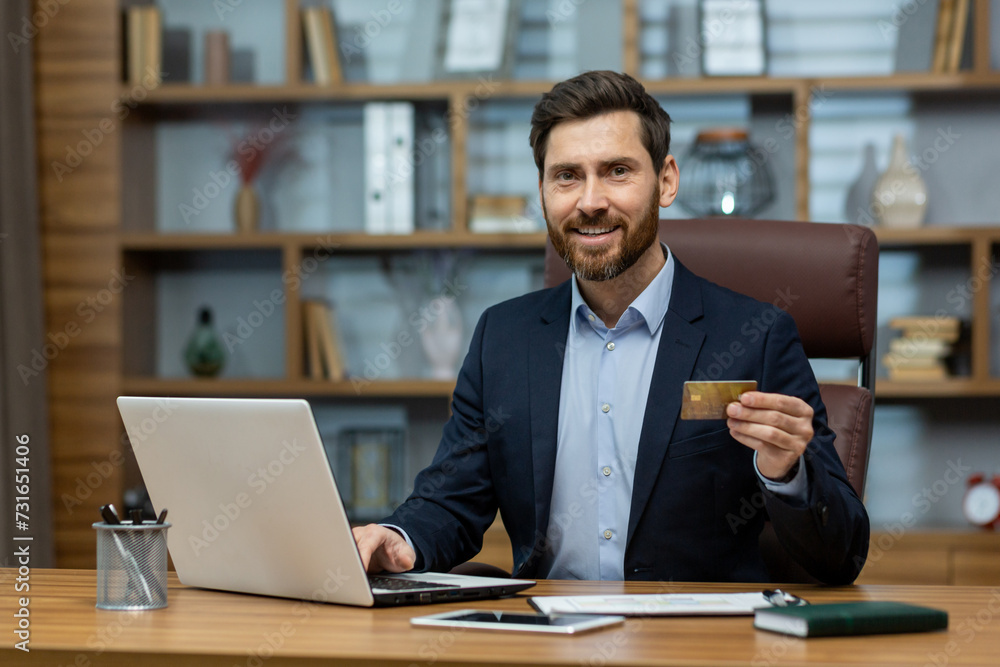 Confident mature businessman in suit with credit card, using laptop at messy home office desk, symbolizing remote work. Confident mature businessman in suit with credit card, using laptop at messy home office desk, symbolizing remote work.