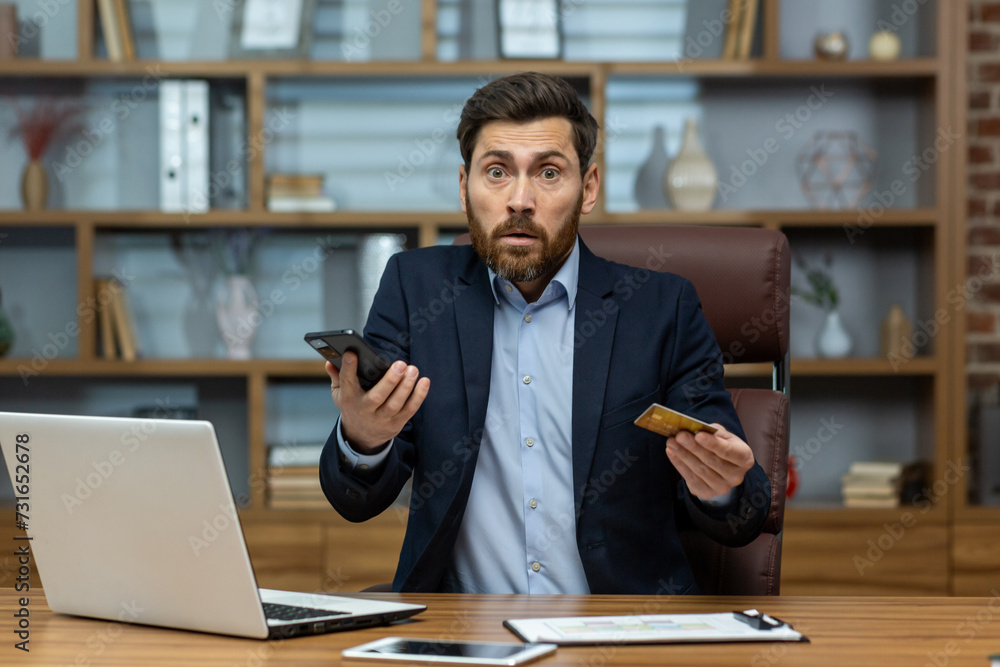 Adult businessman in a suit looks shocked while multitasking in a messy home office, holding a phone and credit card. Adult businessman in a suit looks shocked while multitasking in a messy home office, holding a phone and credit card.
