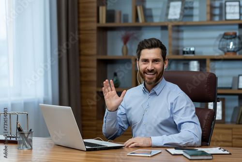 Cheerful mature man in a business suit waving during a video conference in his well-organized home office environment.