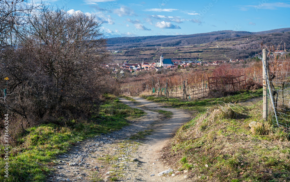 Unpaved road in the hill near vineyards and bushes. View of the church and the small town. In the background there are hills and the horizon. Blue sky with white clouds.