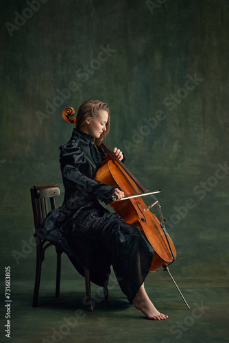 Photos Tender young woman in black dress, cellist sitting on chair and playing cello over dark green background