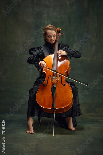 Canvas Print Beautiful young woman, cellist sitting on chair and playing cello against vintage green background