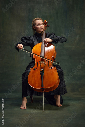 Beautiful woman, talented, passionate cellist looking upward and playing cello against vintage green background. Breathtaking moment. Concept of classical art, retro style, music, inspiration photo