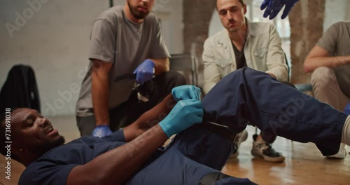 Close-up shooting: A Black male doctor in a blue medical uniform demonstrates the tightening of a medical tourniquet on his leg for public. Practical trainings on first medical aid. Course of a medic photo