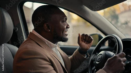 Happy cheerful man Businessman with Black skin in a brown jacket drives a car while sitting in a modern car interior and dances during his business trip in the city photo