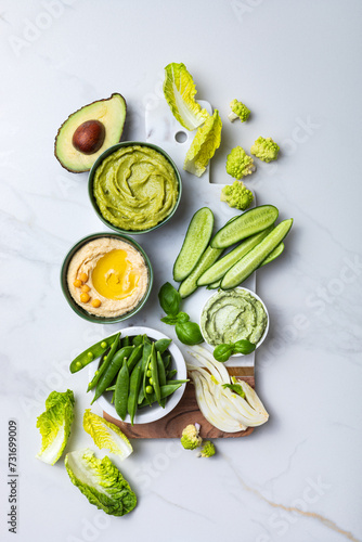Green spring vegetable appetizer platter with dips. Healthy crudites snack board on white marble background.