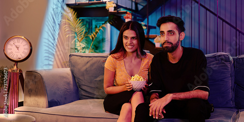 Indian Asian Hindu smiling cute gen z couple young girl boy sitting sofa cozy couch watch movie enjoy indoor home happy adult relax male female pair hold bowl eat snack look fun joy TV film late night