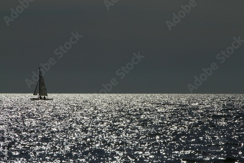 Sailboat is seen cruising across a shimmering body of water, with the sun glinting off the sea