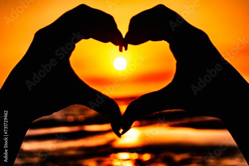 Close-up shot of two hands making a heart shape in front of a bright  golden sunset