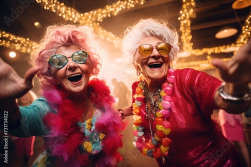 Two senior women with Caucasian features, laughing heartily in a festive setting, adorned with colorful boas, oversized sunglasses, and vibrant beads, exuding happiness and playful energy
