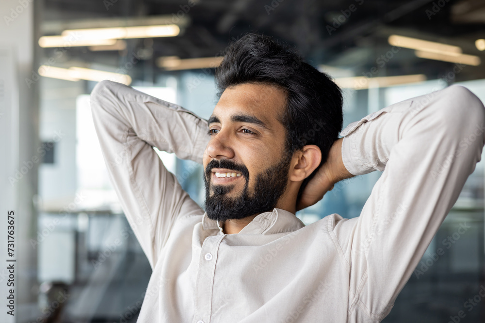 Obraz premium Happy and successful young Indian man, businessman and office worker sitting at workplace with hands behind head and looking ahead. Resting, satisfied with the work and the result. Close-up photo