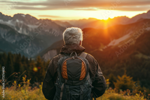 Close up back view of elderly mature man with a travel backpack on his back stands on mountain at sunset. Joyful free travel concept