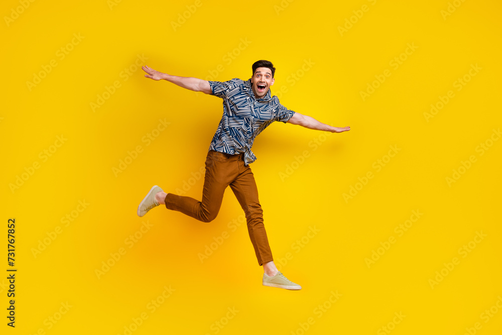 Full length photo of childish funky man dressed print shirt brown trousers flying hold arms like wings isolated on yellow color background Full length photo of childish funky man dressed print shirt brown trousers flying hold arms like wings isolated on yellow color background