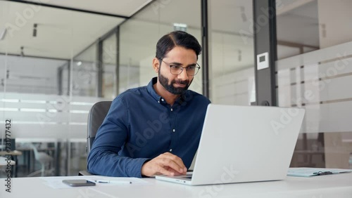 Smiling Indian business man looking at laptop using computer working in office. Busy professional businessman employee or company executive searching online solutions at workplace.