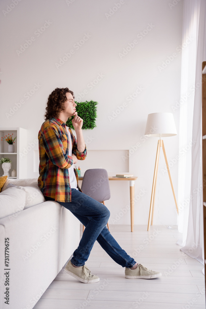 Full length vertical photo of dreamy thoughtful guy wear plaid shirt hand arm chin looking window indoors room home house Full length vertical photo of dreamy thoughtful guy wear plaid shirt hand arm chin looking window indoors room home house