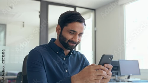 Happy professional businessman employee looking at smartphone checking banking apps typing on cell sitting at desk. Smiling busy Indian business man executive using mobile phone working in office. photo