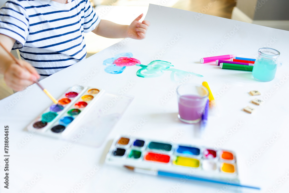 A child in a striped T-shirt dips a brush in watercolor paints. A small child draws on white sheets with paints and markers. Homeschooling, schooling, creative lessons. A child in a striped T-shirt dips a brush in watercolor paints. A small child draws on white sheets with paints and markers. Homeschooling, schooling, creative lessons.