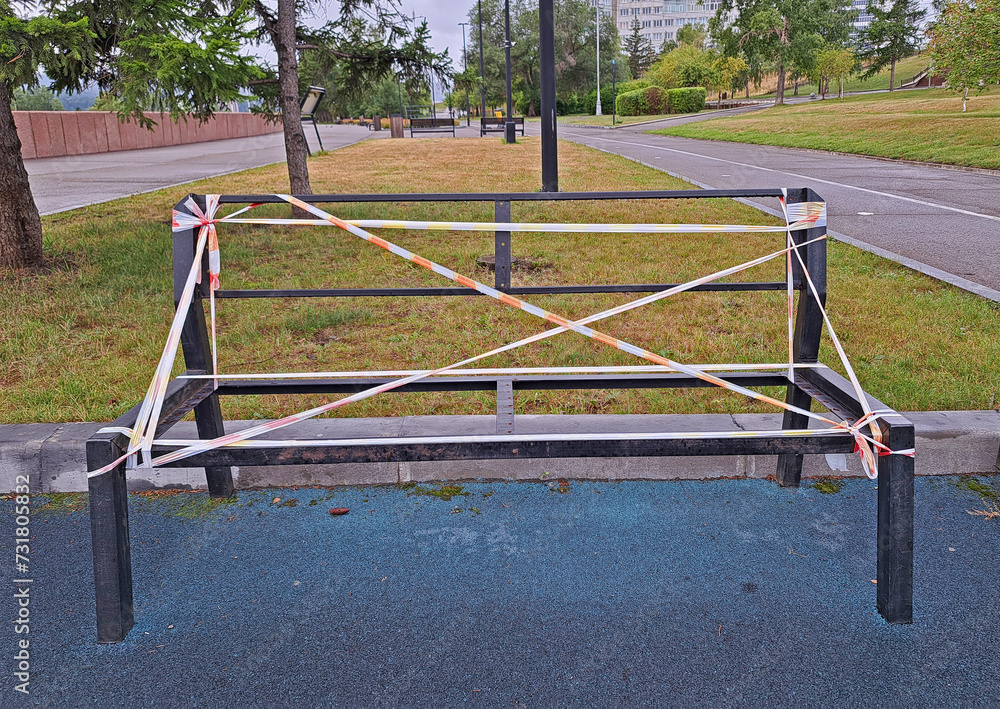 Metal frame of a broken bench. Broken bench in an empty city park.A bench without a seat or backrest is surrounded by warning tape Metal frame of a broken bench. Broken bench in an empty city park.A bench without a seat or backrest is surrounded by warning tape
