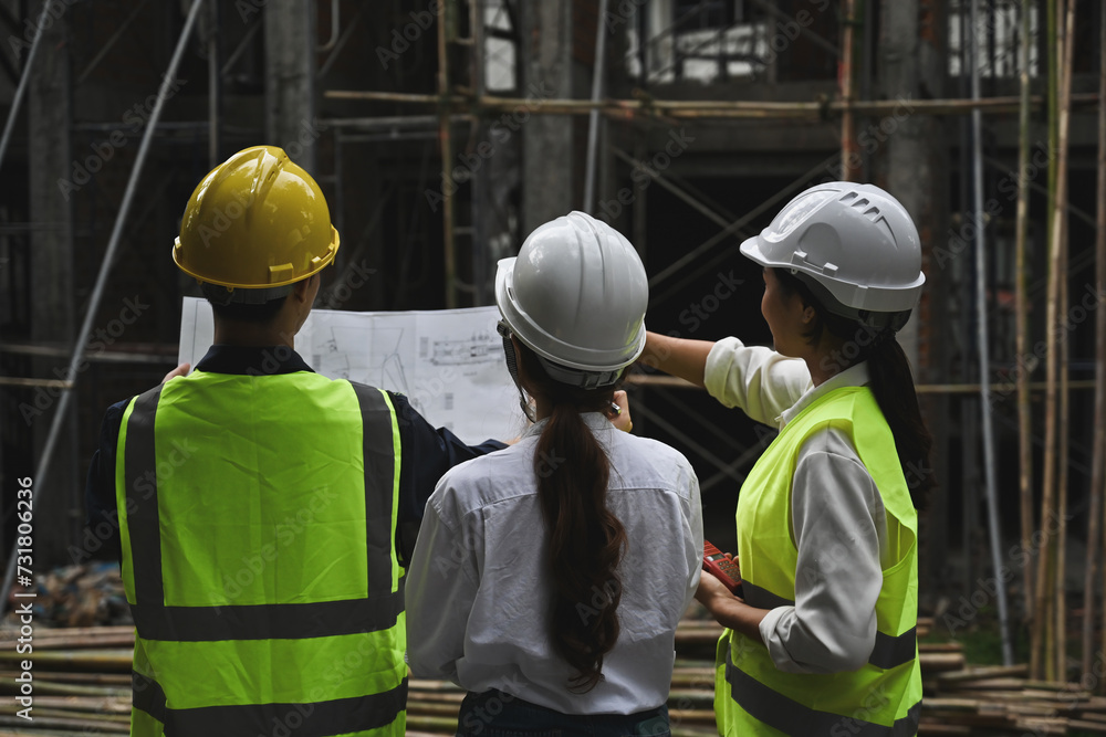 Fototapeta premium Back view of construction project team holding blueprints while inspecting building at construction site