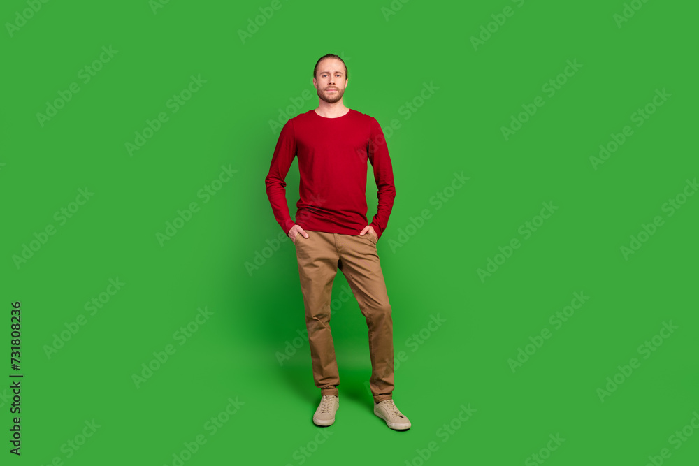 Full length photo of serious handsome guy wear red sweatshirt brown trousers keep palms in pockets isolated on green color background Full length photo of serious handsome guy wear red sweatshirt brown trousers keep palms in pockets isolated on green color background