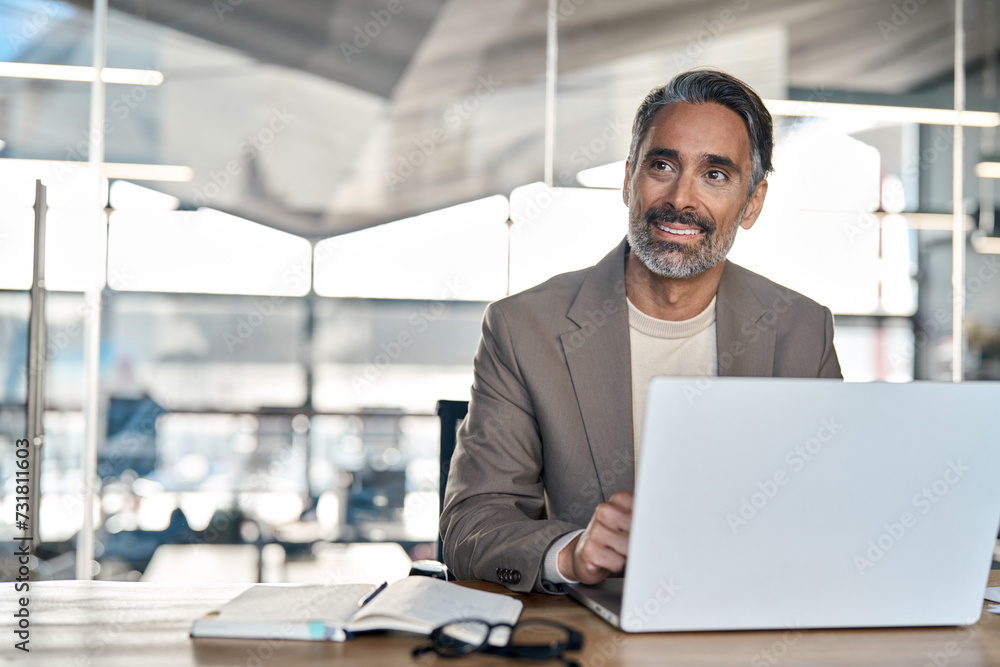 Smiling mature business man executive wearing suit sitting at desk using laptop. Happy busy professional middle aged businessman investor banker working on computer looking away in office. Copy space. Smiling mature business man executive wearing suit sitting at desk using laptop. Happy busy professional middle aged businessman investor banker working on computer looking away in office. Copy space.