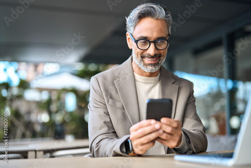Happy stylish older busy businessman investor holding smartphone looking at cellphone doing banking payments. Middle aged business man using mobile cell phone working outdoors. Candid photo.