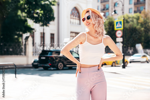 Young beautiful smiling blond hipster woman in trendy summer clothes. Carefree female posing in the street at sunny day. Positive model outdoors at sunset. Cheerful and happy in hat, sunglasses