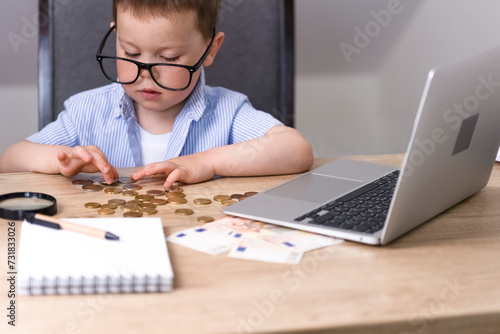 A little boy in a blue shirt and glasses counts money, euros, cents. The child counts money, there is a magnifying glass on the table, there is a laptop and there are papers. High quality photo