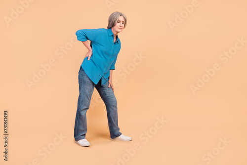 Full length photo of tired suffering pensioner person wear blue shirt jeans hold hand on lower back isolated on beige color background