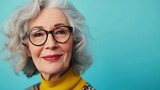 Elegant elderly woman with gray hair wearing glasses smiling and dressed in a yellow turtleneck and patterned top against a blue background.