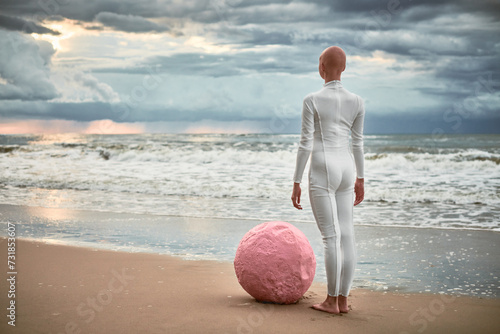 Hairless performer girl with alopecia in white futuristic suit stands on sea beach with pink sphere at restless clouds sky, surreal scene exudes confidence and hope resilience against life challenges