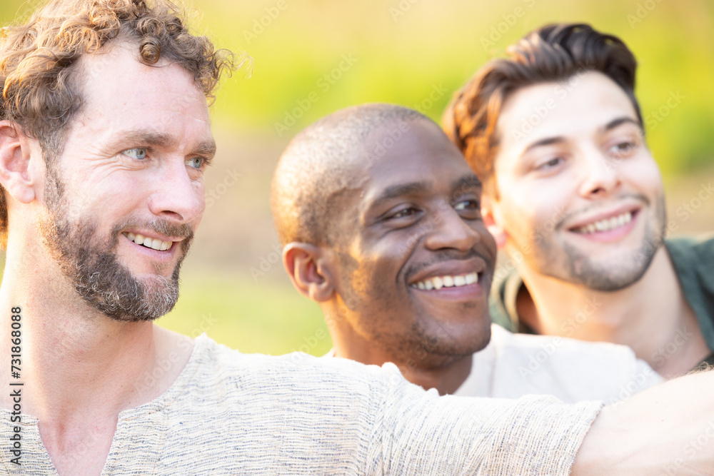 This image portrays a group of three men of different ethnicities sharing a joyful moment outdoors. The man in the foreground, with curly hair and a light stubble, is looking away with a smile, as if This image portrays a group of three men of different ethnicities sharing a joyful moment outdoors. The man in the foreground, with curly hair and a light stubble, is looking away with a smile, as if