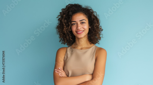 woman with curly hair is smiling at the camera, standing with her arms crossed in front of a light blue background