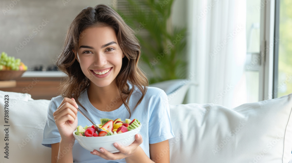 A cheerful woman sitting on a sofa, eating a fresh and colorful salad from a bowl, looking happy and healthy.