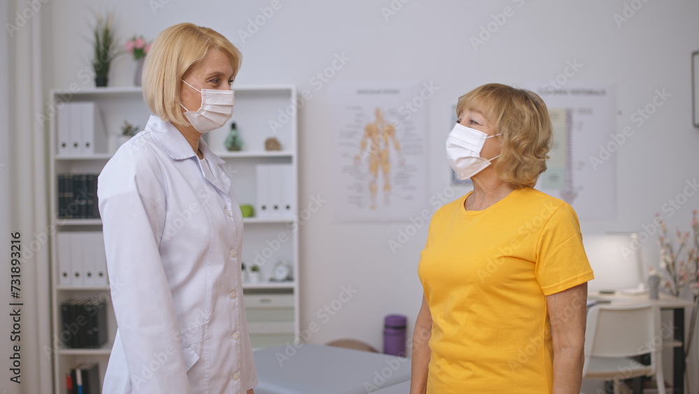 Fototapeta premium A positive female doctor and a senior patient, both in face masks, look at each other optimistically, celebrating recovery