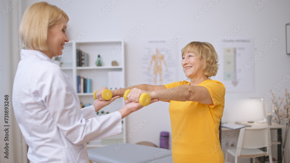 An excited patient and doctor exercise with dumbbells together at a rehabilitation clinic An excited patient and doctor exercise with dumbbells together at a rehabilitation clinic