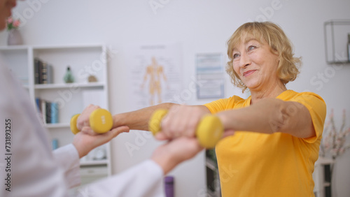 A positive mature lady exercises with dumbbells, supported and assisted by her doctor
