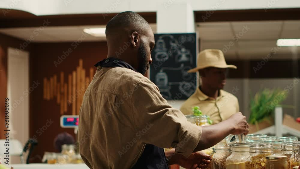 Merchant with apron placing bulk items on shelves at eco store, supporting zero waste concept and sustainable lifestyle. African american vendor advertising homemade pantry supplies. Camera A. Merchant with apron placing bulk items on shelves at eco store, supporting zero waste concept and sustainable lifestyle. African american vendor advertising homemade pantry supplies. Camera A.