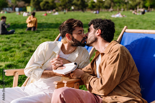Cuadro en lienzo Loving gay couple sitting on deckchair and kissing in park