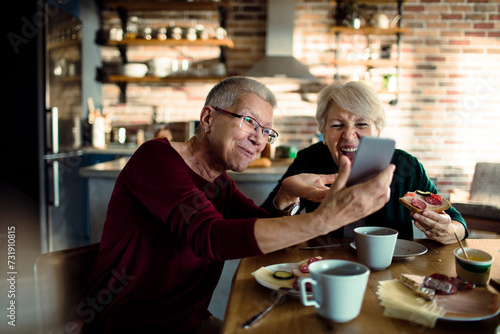 Two senior women eating breakfast with a smartphone in the kitchen