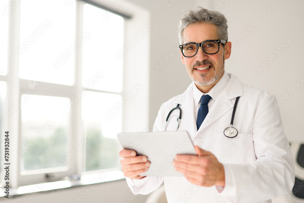 Senior doctor with glasses and a friendly demeanor holds a tablet, standing by a window with natural light that suggests a modern and approachable healthcare setting. Contemporary medical practice Senior doctor with glasses and a friendly demeanor holds a tablet, standing by a window with natural light that suggests a modern and approachable healthcare setting. Contemporary medical practice