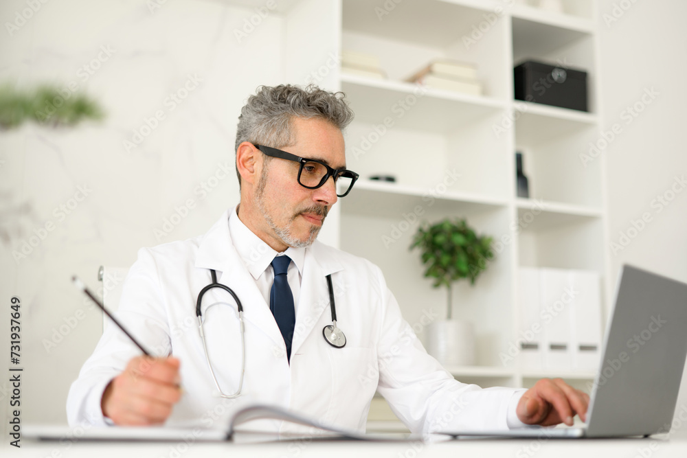 Focused senior doctor with grey hair reviews patient information on a laptop and taking notes, symbolizing the integration of technology in modern medical practice, within contemporary clinic setting Focused senior doctor with grey hair reviews patient information on a laptop and taking notes, symbolizing the integration of technology in modern medical practice, within contemporary clinic setting